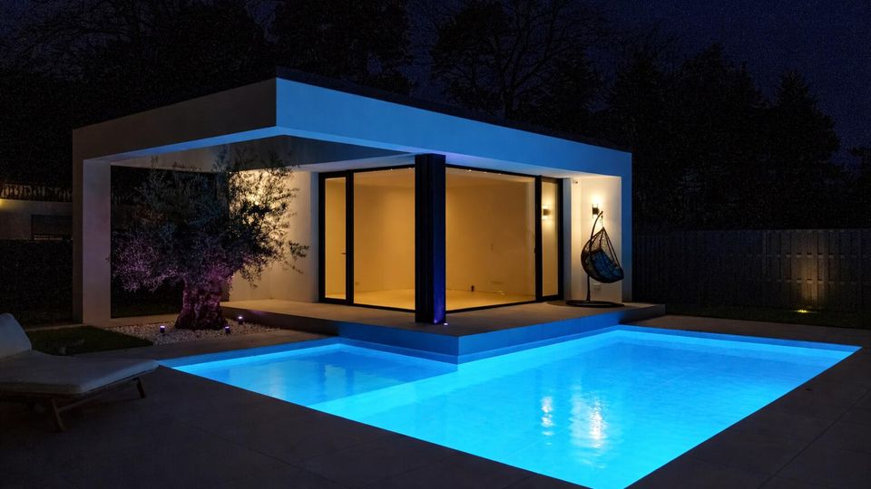 Modernes, flaches Pavillongebäude mit breitem Vordach und großen bodentiefen Glasfronten, nachts warm beleuchtet. Davor liegt ein rechteckiger, blau beleuchteter Swimmingpool mit umlaufender Terrasse; im Hintergrund sind Bäume und ein Sichtschutzzaun zu sehen.