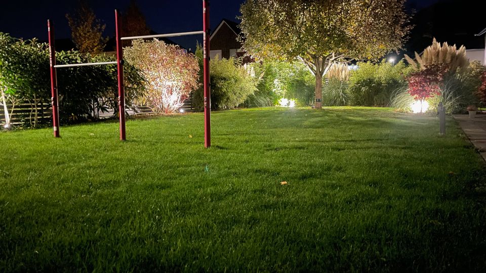 Grünfläche eines Gartens bei Nacht, beleuchtet durch mehrere Bodenlichter. Im Vordergrund steht eine rot-weiße Turnstange, im Hintergrund sind Büsche, Ziergräser und ein einzelner Baum zu sehen.