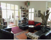 Wohnzimmer mit schwarzen Ledersofas, roten Kissen und orientalischem Holztisch auf bunten Teppichen. Im Hintergrund gläserne Doppeltüren, die zu einem hellen Wintergarten mit weißen Möbeln und dekorativen Statuen führen.