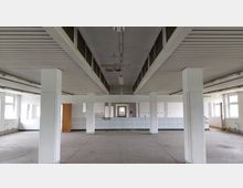 Ein leerer, rechteckiger Innenraum mit weißen Wänden und mehreren Stützpfeilern. Die Decke besteht aus paneelartigen Elementen mit eingelassenen Leuchten, die Wände sind teilweise mit weißen Fliesen verkleidet, und am hinteren Ende befinden sich ein Durchgang und eine Holztür.