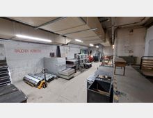 Industriehalle mit Lagerbereich, ausgestattet mit gestapelten Materialien wie Metallplatten, Rohren und Werkzeugen. An einer Betonwand steht in roter Schrift „RAUCHEN VERBOTEN“, und im Hintergrund sind Treppen sowie zusätzliche gelagerte Gegenstände sichtbar.