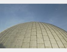 Kuppelförmiges Gebäude mit einer Fassade aus gleichmäßig angeordneten, transparenten Paneelen, fotografiert vor einem klaren blauen Himmel.