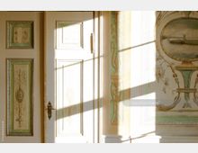 Detail einer historischen Innenraumtür mit klassizistischen Verzierungen in Cremeweiß und Pastellgrün. Die Wand daneben ist mit kunstvollen Malereien wie floralem Dekor und einem Landschaftsmedaillon gestaltet; einfallendes Sonnenlicht wirft Schatten auf die Oberfläche.