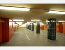 Unterirdischer Durchgang mit gelben, roten und grünen gefliesten Wänden, unterstützt durch blau-grüne Säulen. Die Decke ist beige, und mehrere Deckenleuchten sorgen für eine gleichmäßige Beleuchtung.