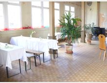 Ein einfacher Innenraum eines Gastraums mit weißen Tischdecken gedeckten Tischen, Holzbänken und -stühlen, sowie mehreren großen Zimmerpflanzen in Töpfen. Der Raum hat hohe Fenster mit farbigen Stoffen auf den Fensterbänken und wirkt leicht renovierungsbedürftig.