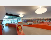 Innenansicht einer modernen Kinderbücherei mit orangefarbenen, geschwungenen Regalen, die mit Büchern gefüllt sind. Große Fensterfront mit Blick nach draußen, Sitzsäcke und niedrige Sitzbereiche auf dem hellen Boden; zwei große, runde Deckenleuchten erhellen den Raum.