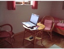 Ein kleines Zimmer mit Holzboden, einem runden Rattantisch mit aufgeklapptem Laptop darauf und zwei passenden Rattanstühlen mit rot-weiß karierten Sitzkissen. Im Hintergrund befindet sich ein Bett mit einer rot-weiß karierten Bettwäsche und ein Fenster mit gleicher Gardine.