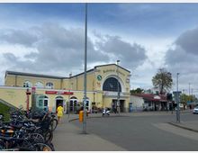 Bahnhofsgebäude in Erkner mit gelber Fassade, großen Rundbogenfenstern und Schriftzug „Bahnhof Erkner“. Im Vordergrund befinden sich Fahrräder, eine Fußgängerin, sowie kleinere Läden und ein Café seitlich des Gebäudes.
