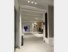 Ein modernes, minimalistisch eingerichtetes Wohnzimmer mit weißen Wänden und einer weißen Decke mit sichtbaren Balken. Im Raum befinden sich abstrakte Kunstwerke an den Wänden, ein heller Teppich, eine Vase mit Blumen und eine Marmoroberfläche in der rechten Ecke.