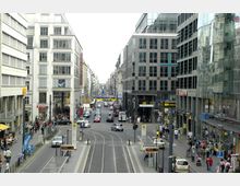 Straßenszene in einer Stadt mit moderner Architektur; links und rechts stehen mehrstöckige Gebäude mit Glasfassaden und Geschäften im Erdgeschoss. Die Straße hat Straßenbahnschienen in der Mitte, daneben fahren Autos, während Gehwege mit Passanten belebt sind.
