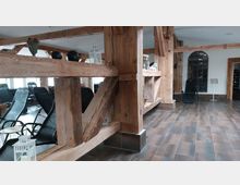 Innenraum eines rustikalen Entspannungsbereichs mit sichtbaren Holzbalken, braunen Fliesen und Liegestühlen. Im Hintergrund sind dekorative Elemente wie Laternen und ein großes rundes Fenster zu sehen.