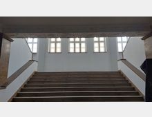 Breite Holztreppe in einem Gebäude mit hellem Innenraum, flankiert von weißen Wänden und schlichten Holzgeländern, führt zu einer Wand mit großen Sprossenfenstern, die diffuses Tageslicht hereinlassen.