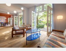 Wohnraum mit Holzfußboden, großen Fenstern und offenen Glastüren, die zu einem Garten führen. Im Vordergrund stehen farbenfrohe Sitzmöbel und ein blauer Glastisch, im Hintergrund ist ein Essbereich mit orangefarbener Wand zu sehen.