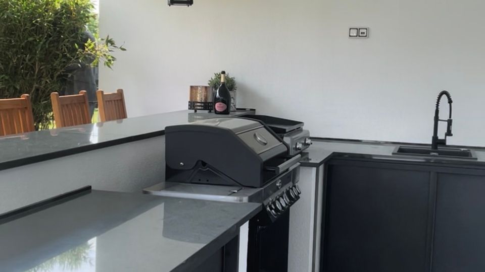 Überdachte Außenküche auf einer Terrasse mit weißen Deckenbalken und hängenden Laternenleuchten, ausgestattet mit dunklen Unterschränken und grauer Arbeitsplatte. Im Vordergrund stehen ein Gasgrill und rechts eine Spüle; im Hintergrund sind Pflanzen und Sitzmöbel zu sehen.