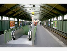 Überdachter Bahnsteig einer U-Bahn-Station mit einem grünen Metallgerüst und Fenstern entlang der oberen Wand. In der Mitte befindet sich eine Rolltreppe, umgeben von einem Geländer, und im Hintergrund sind Fahrgäste, Sitzbänke sowie Züge zu sehen.