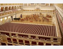 Innenansicht eines Konzertsaals mit roten Sitzreihen und goldverzierter weißer Balustrade. Im Vordergrund befinden sich leere Sitzplätze, während auf der Bühne ein Flügel und Instrumentenstühle für ein Orchester angeordnet sind.