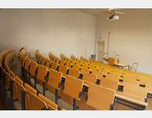Ein Hörsaal mit ansteigenden Reihen aus hölzernen Klappsitzen, einem Dozentenpult mit Mikrofon, zwei Tischen und einer Deckenprojektorinstallation. Die Wände sind schlicht, und hinter dem Pult befinden sich Türen und ein Papierkorb.