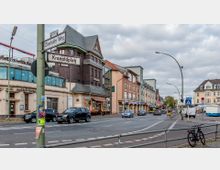 Straßenszene mit belebter Kreuzung, umgeben von mehrstöckigen Gebäuden mit Geschäften und Restaurants im Erdgeschoss. Ein Schild weist auf den "Oberhofer Weg" und den "Kranoldplatz" hin, Autos und Fahrräder prägen das Stadtbild, während im Hintergrund ein Wochenmarkt zu sehen ist.