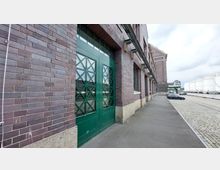 Backsteinfassade eines Industriegebäudes mit großen grünen Doppeltüren und Fenstern mit Gittermuster. Rechts verläuft ein gepflasterter Weg entlang der Gebäude, im Hintergrund sind Industrieanlagen und geparkte Autos erkennbar.