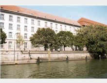 Großes, vierstöckiges Gebäude mit heller Fassade und rotem Satteldach, gelegen an einem Fluss mit einer steinernen Uferbefestigung. Mehrere Bäume stehen vor dem Gebäude, das von einem ruhigen Gewässer umgeben ist.