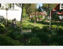 Ein gepflegter Garten mit grünen Rasenflächen, niedrigen Sträuchern, Bäumen und Blumenbeeten, in dem eine Skulptur auf einem Sockel steht. Im Hintergrund sind Gebäude mit roten Ziegeldächern und weißer Fassade sichtbar.