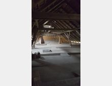 Dachboden eines Gebäudes mit offenem Holzgebälk und schrägem Dach. Der Raum ist unrenoviert, mit freiliegenden Balken, Holzböden und vereinzelten Baustoffen.