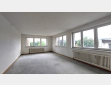 Leerer Raum mit weißen Wänden, großem Fensterbereich auf zwei Seiten und Heizkörpern darunter. Der Boden ist unbehandelt, und das Fenster bietet Blick auf eine grüne Umgebung und benachbarte Gebäude.