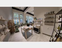 Ein kleiner, heller Laden mit großen Fenstern und hohen Decken; Regale mit Keramiktassen und Weinflaschen sowie Tische mit Büchern und Pflanzen. Auf der linken Seite ein geflochtener Regalständer und weitere Dekorationsartikel.