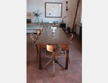 Ein schlicht eingerichteter Raum mit einem großen rechteckigen Holztisch und sieben Stühlen, umgeben von neutralen Wänden. Im Hintergrund stehen ein Regal mit Büchern und Glasgeschirr, ein gerahmtes Bild an der Wand sowie eine Leiter, die an der rechten Seite lehnt.