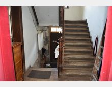 Holztreppe in einem älteren, schlichten Gebäude mit sichtbaren Gebrauchsspuren. Links ein Heizkörper an der Wand, rechts ein Seilabsperrung, und im unteren Bereich eine Holztür mit Glasfenster.