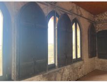 Ein Innenraum mit alten, dunklen Spitzbogenfenstern, die teilweise mit Holzläden verschlossen sind. Die Wände bestehen aus groben, unverputzten Ziegelsteinen und weisen sichtbare Abnutzung auf.