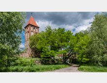 Historischer Kirchturm aus rotem Backstein mit spitzem Dach, umgeben von grünen Bäumen und Grasflächen unter einem bewölkten Himmel. Im Vordergrund führt ein unbefestigter Weg durch die Landschaft.