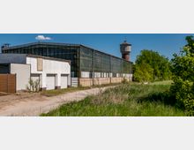 Industriegebäude mit Glasfassade und angrenzendem weißen Anbau mit Garagentoren, daneben ein alter Wasserturm aus Backstein. Im Vordergrund sind eine unbefestigte Straße und Wiese, im Hintergrund stehen Bäume unter klarem Himmel.