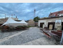 Gepflasterter Hof mit Sitzbänken und Tischen, teilweise überdacht mit einem weißen Segeltuch. Im Hintergrund sind alte Steinmauern, ein Gebäude mit roten Dachziegeln, ein Funkmast und weitere Gebäude zu sehen.