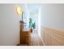 Heller, schmaler Flur mit weiß gestrichenen Wänden und halbhoher, cremefarbener Holzvertäfelung. Links steht eine Holzkommode mit dekorativen Vasen, darüber ein runder Spiegel und eine Wandleuchte. Am Ende des Flurs stehen große grüne Zimmerpflanzen in Töpfen, und eine geöffnete Tür führt in einen weiteren Raum.