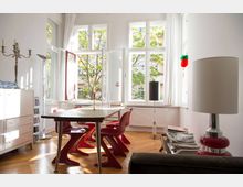 Helles Wohnzimmer mit großen, weißen Fenstern, durch die Tageslicht hereinfällt, und einer Aussicht auf Bäume. Es ist mit einem Holztisch, roten Designstühlen, einem weißen Sideboard, einer Stehlampe und einem Kronleuchter ausgestattet; der Boden besteht aus Holzparkett.