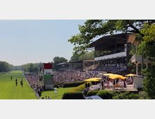 Eine Pferderennbahn mit einer langen, grünen Rennstrecke, flankiert von dicht gedrängten Zuschauern. Rechts im Bild befinden sich überdachte Zuschauertribünen und ein Bereich mit gelben Sonnenschirmen, während Bäume den Hintergrund säumen.
