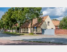 Straßenszene mit einem historischen, einstöckigen Wohnhaus mit Satteldach und hellbeiger Fassade, umgeben von Bäumen und niedrigen Zäunen. Im Vordergrund führt eine asphaltierte Straße vorbei, flankiert von einem Gehweg aus Pflastersteinen.