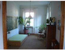 Schlicht eingerichtetes Schlafzimmer mit einem niedrigen Holzbett und grüner Bettdecke, mehreren Zimmerpflanzen, einem geflochtenen Stuhl und einem hölzernen Bücherregal. Ein Fenster mit weißen Vorhängen lässt Tageslicht herein, und eine hängende Lampe beleuchtet den Raum.