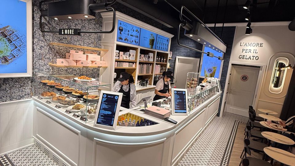 Innenansicht einer modernen Eisdiele mit einer geschwungenen, weißen Theke, auf der Speisen und Getränke präsentiert werden. Der Raum ist mit dekorativen Fliesenboden, hängenden Lampen und Sitzbereichen mit kleinen Tischen und schwarzen Stühlen ausgestattet.