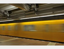 Gelbe U-Bahn, die durch eine unterirdische Station fährt, mit modernem, metallisch gestaltetem Deckenbereich und gefliesten Bodenflächen.