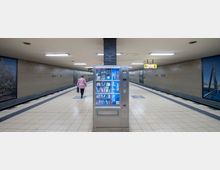 Ein unterirdischer U-Bahnhof mit dem Namen "Britz-Süd". Im Vordergrund steht ein beleuchteter Snack- und Getränkeautomat, im Hintergrund laufen Menschen entlang des hell gefliesten Bodens, flankiert von beige gekachelten Wänden mit dekorativen Bildern.