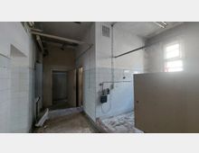 Ein leerer, verlassener Innenraum mit weißen, teilweise beschädigten Fliesen an den Wänden, freiliegenden Rohren an der Decke und einem offenen Türrahmen im Hintergrund. Der Boden ist uneben und teilweise verschmutzt, während ein vergittertes Fenster schwaches Tageslicht hereinlässt.