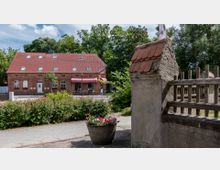 Backsteinbau mit rotem Satteldach, großen Fenstern und einer Markise mit der Aufschrift „Kaffee, Kuchen, Eis“, umgeben von Bäumen. Im Vordergrund ein Blumenkübel und eine Mauer mit Holzzaun.