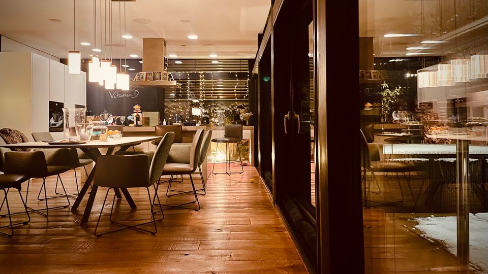 Modernes Café- oder Lounge-Interieur mit Holzfußboden, großem Esstisch und gepolsterten Stühlen unter hängenden Pendelleuchten. Rechts verläuft eine dunkle Glasfront mit Blick auf eine beleuchtete Theke und Regale im Hintergrund.