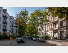 Straßenszene in einer Wohngegend mit Reihen von Altbauwohnungen auf beiden Seiten; links weiße Fassaden mit Balkonen und rechts ältere Gebäude mit grauen, teils beschädigten Fassaden. Es gibt geparkte Autos entlang der Straße, Bäume spenden Schatten, und ein orangefarbener Briefkasten steht auf dem Gehweg.