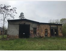 Altes, einstöckiges Backsteingebäude mit verwittertem Dach, großen Holztoren und teilweise beschädigten Fenstern. Im Vordergrund ist eine grasbewachsene Fläche, im Hintergrund befinden sich weitere Bäume und ein Haus.