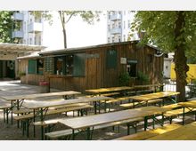 Holzverteilter Biergarten mit langen gelben Holzbänken und Tischen auf einem Kiesboden, umgeben von Bäumen. Im Hintergrund befindet sich ein rustikaler Verkaufsstand mit grünen Fensterläden, hinter dem moderne Wohngebäude sichtbar sind.