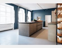 Moderne Küche mit einer großen Kücheninsel aus Betonoptik in der Mitte, blauen Wänden und großen Fenstern mit weißen Rahmen. Im Hintergrund befinden sich eine weiße Tür, ein Wandregal mit Geschirr und eine stilvolle Wanddekoration.