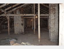 Dachboden mit freiliegendem Holzbalkenwerk, Betonwänden und sichtbarem Dachgebälk. Der Raum wirkt roh und unbenutzt, mit verstreuten Gegenständen und einer schwachen Deckenbeleuchtung.